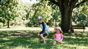 A young boy and girl sitting together outdoors wearing colorful wide-brimmed hats, smiling under bright sunshine on a green lawn.